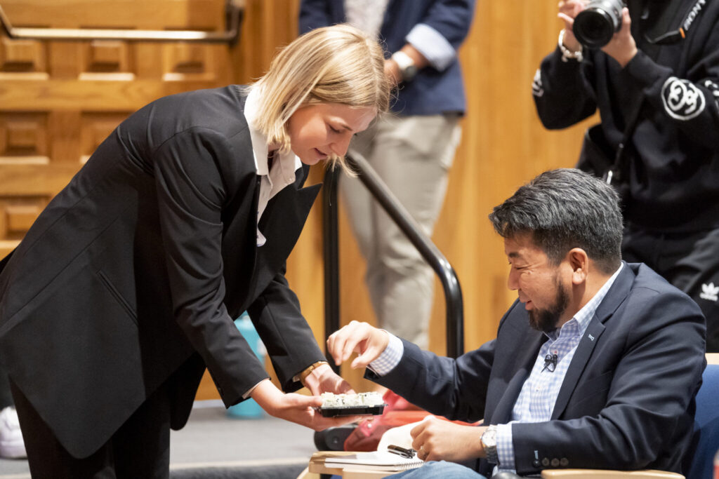 Kate Sullivan holds out a plate of plant-based sushi across a table, which judge Jay Onda reaches towards. Jay is seated at the table.