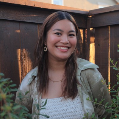 Katrina Manaloto, a young woman, stands in front of a fenced wall with plants at her sides.