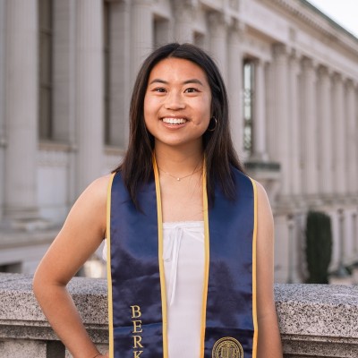 Claire Chu, a young woman, stands smiling in front of UC Berkeley's Doe Library. The library is a large building with columns lining the front. Claire is wearing a blue-and-yellow Berkeley graduation stole.