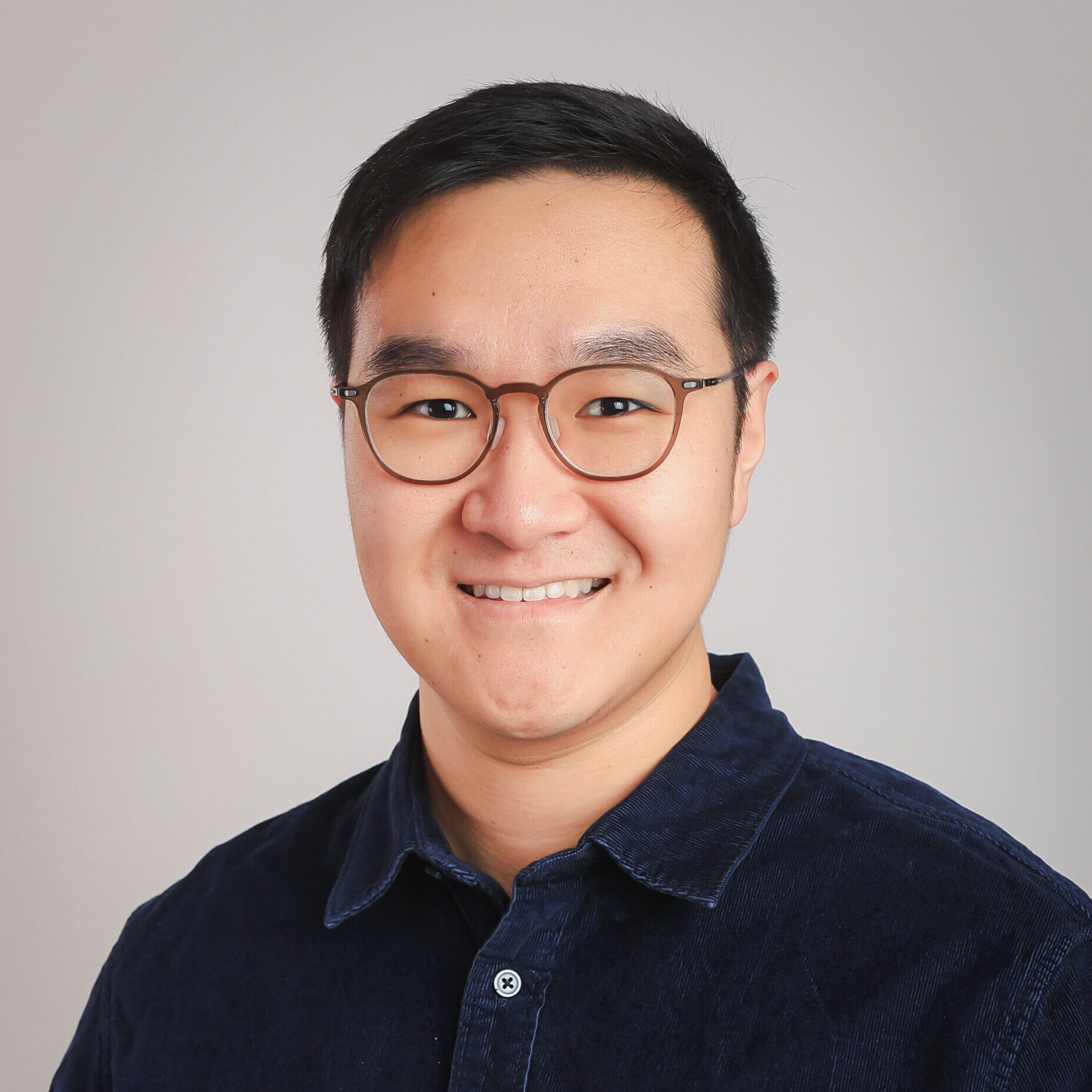 Dylan Chiu smiles for a profesional headshot against a light gray background. He is wearing a black collared shirt.