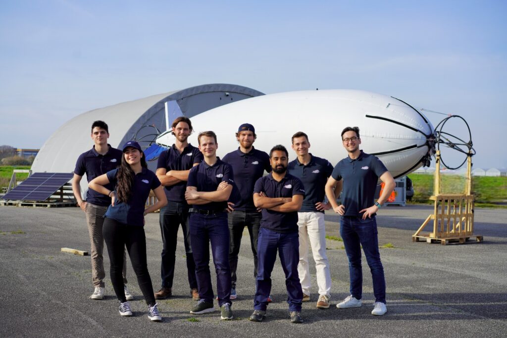 The HyLight team poses in front of the HyLighter, their hydrogen-powered airship drone outside in a natural environment