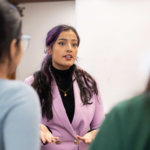 Kashish Juneja, a woman in a pink suit, stands centered and speaking in the direction of the camera. The backs of two students, are seen at the edges of the frame as they listen to Kashish.