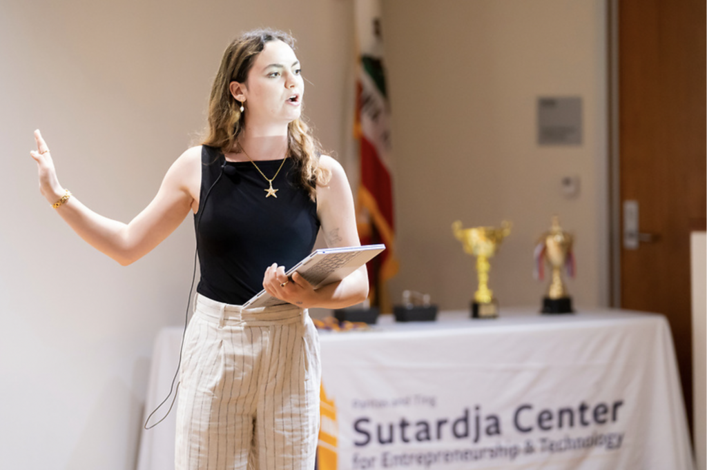 Rachel Eizler gestures while presenting on the Collider Cup stage with trophies on a table in the background.