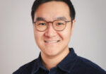 Dylan Chiu smiles for a profesional headshot against a light gray background. He is wearing a black collared shirt.