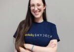 Mar Navarro smiles for a professional photo, wearing a navy blue Berkeley SkyDeck t-shirt against a dark gray wall.