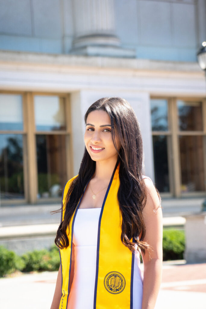 Rashmi Varma poses in front of Doe Memorial Library wearing a gold graduation stole.